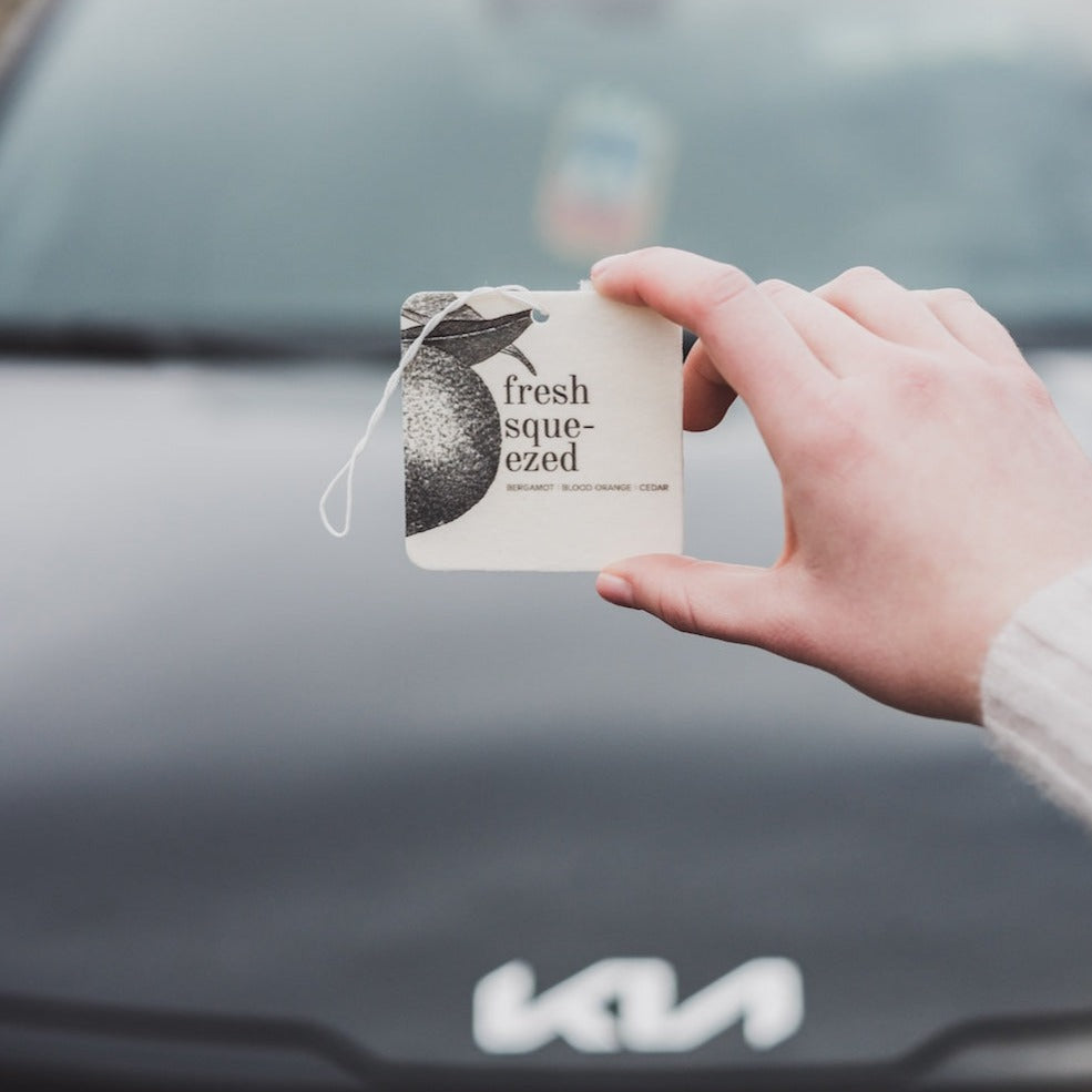 Fresh Squeezed Car Freshener being held in front of a car.