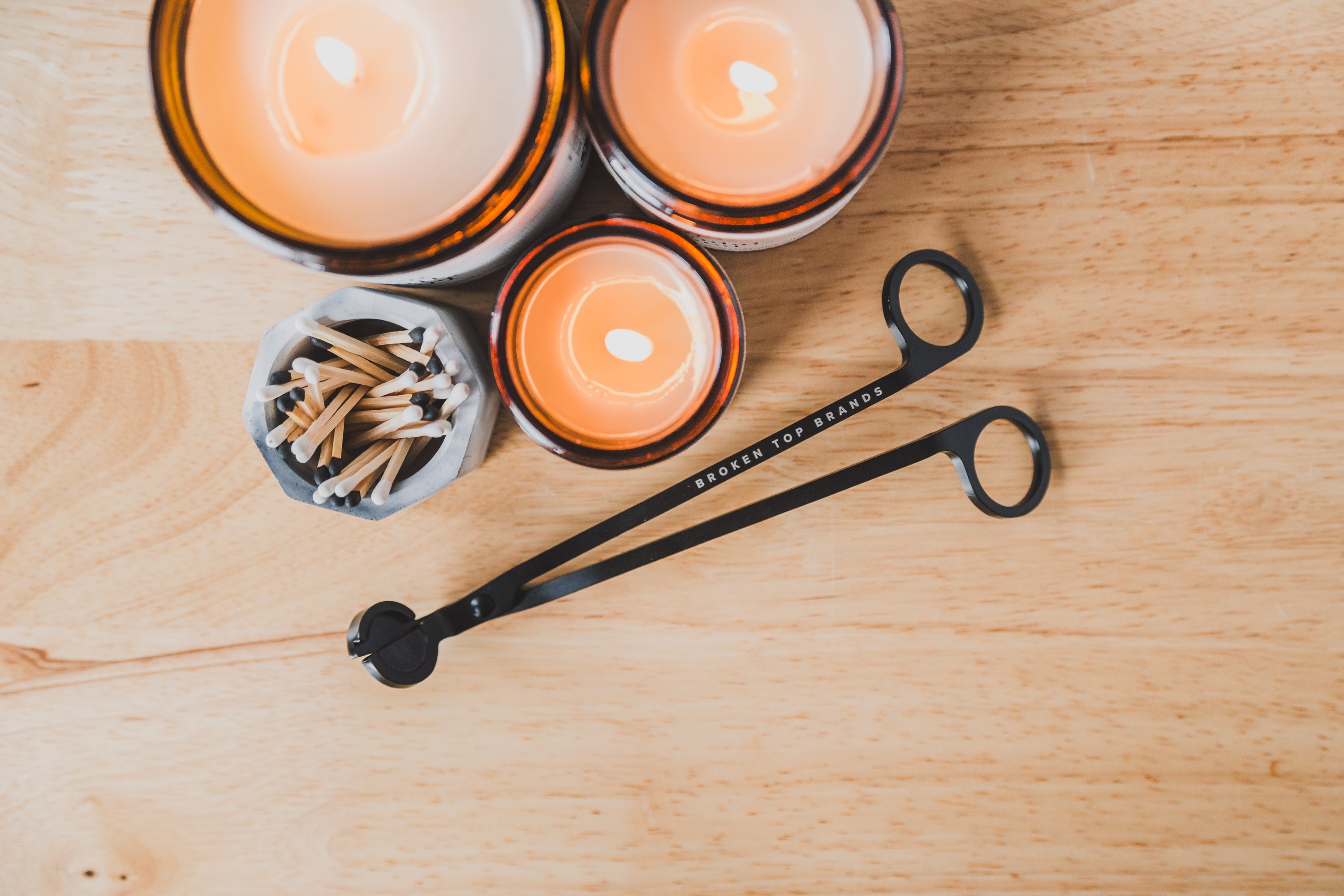 Black Broken Top Brands wick trimmer on wood table next to 3 candles and matches