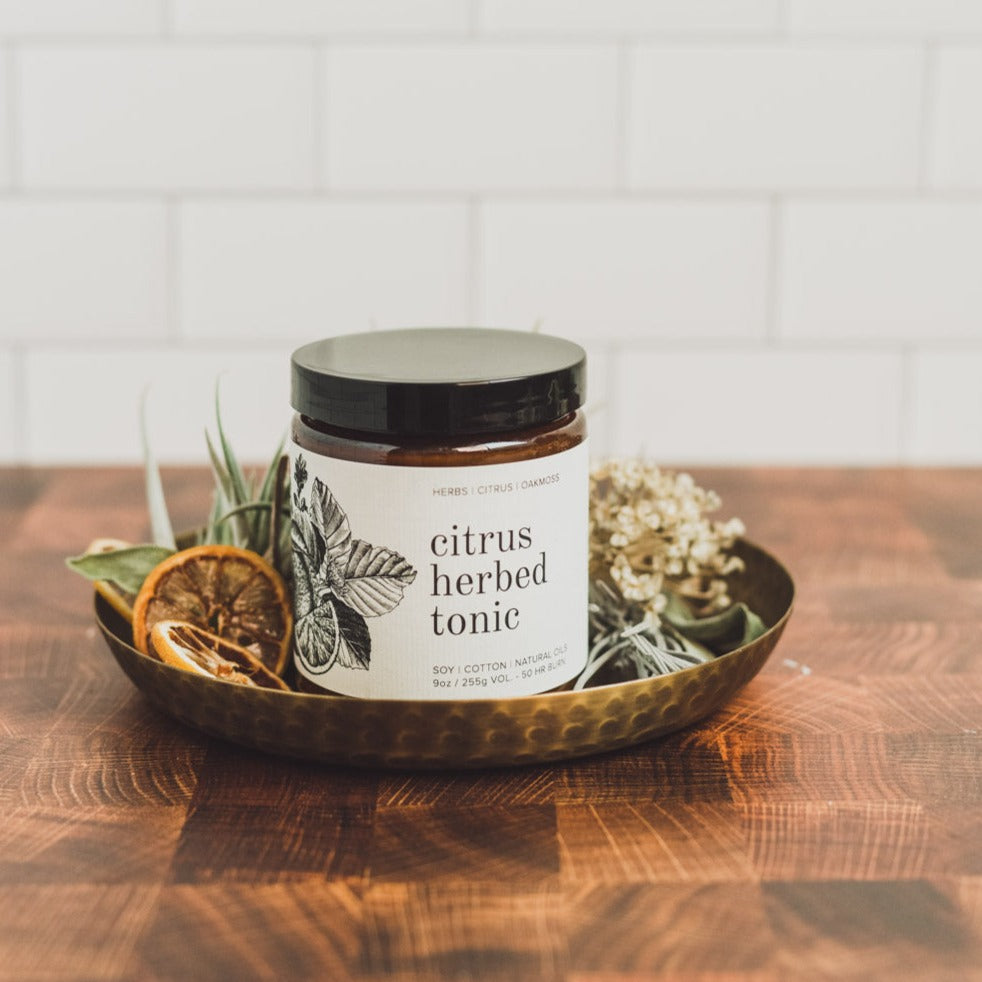 9oz Citrus Herbed Tonic candle on wooden counter surrounded by foliage
