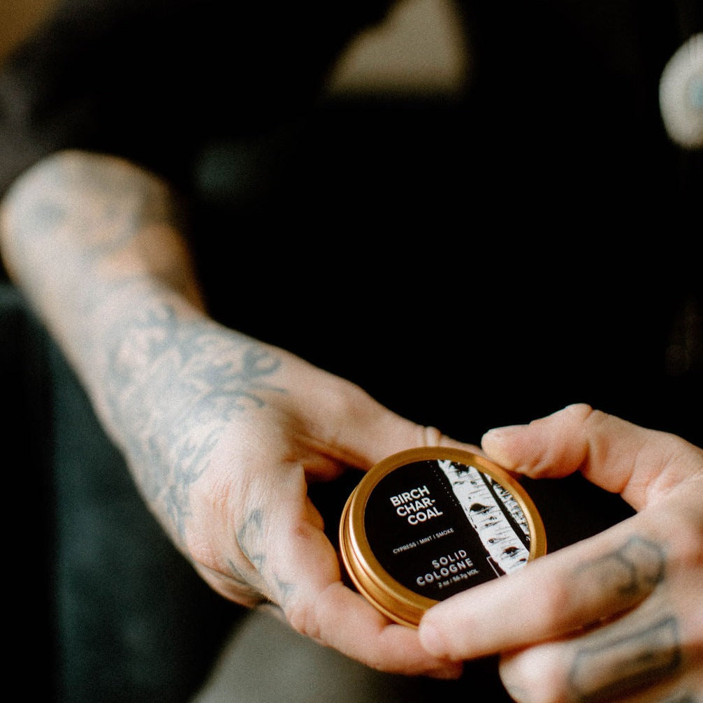 Birch Charcoal solid cologne being held by two tattooed hands