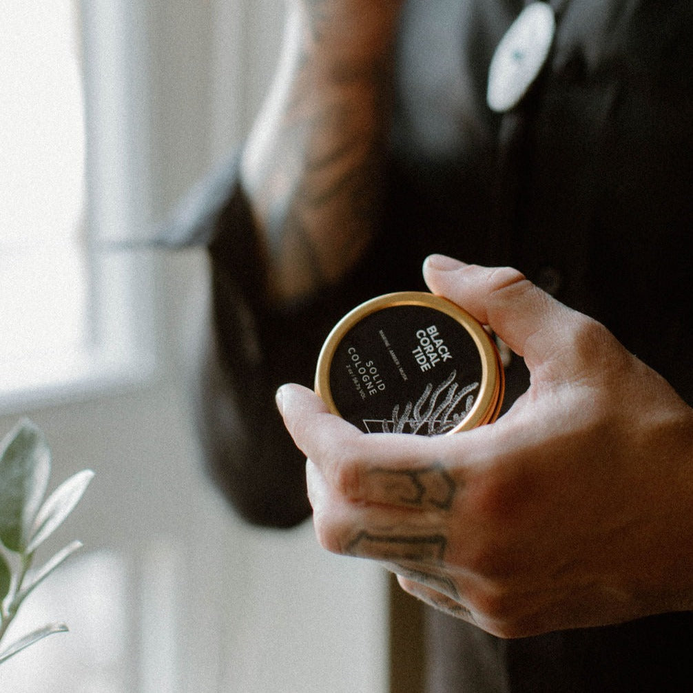 Black Coral Tide solid cologne being held by tattooed hand
