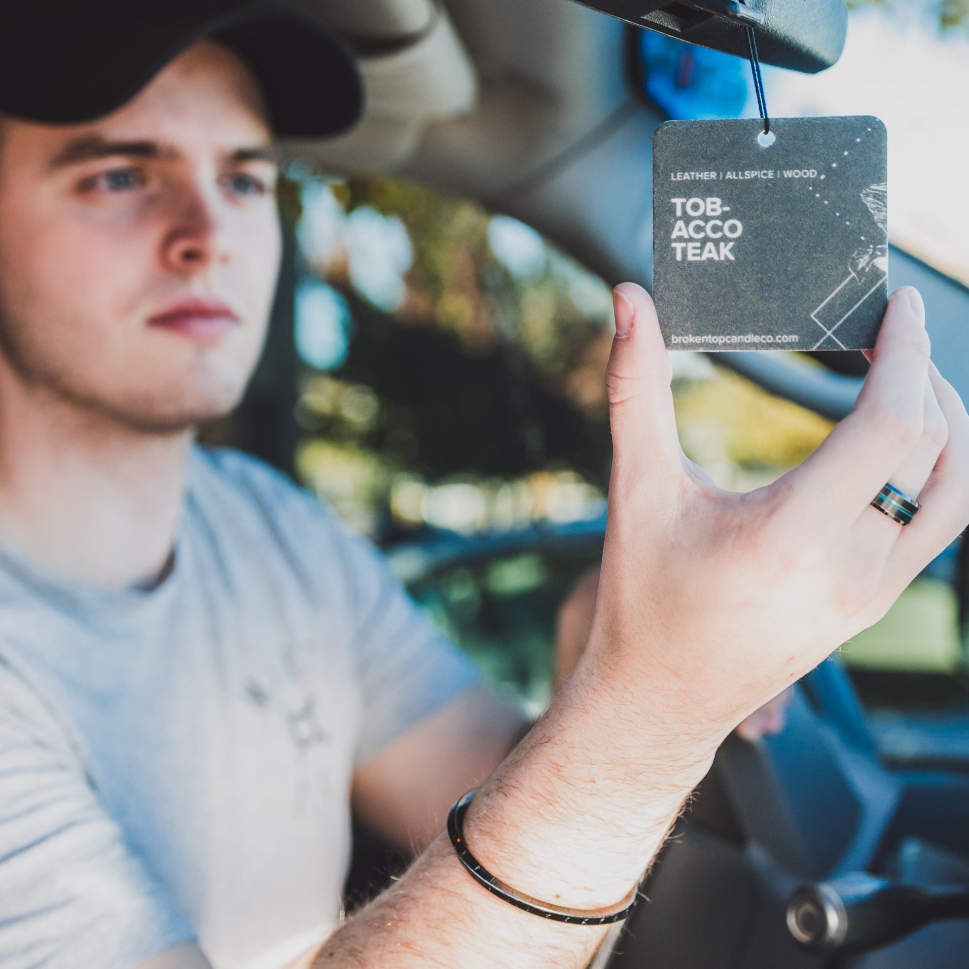 Tobacco Teak car freshener hanging from car rear view mirror with man holding in his hand