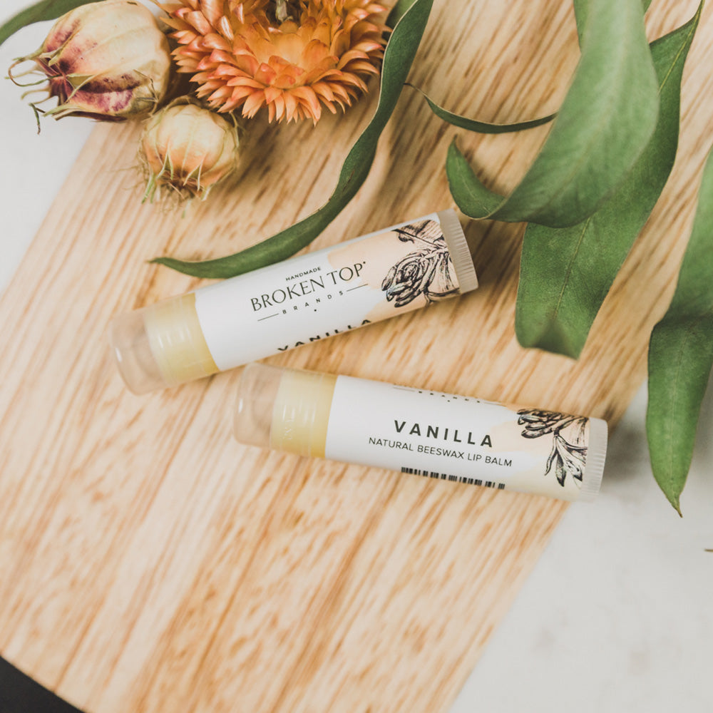 Two Vanilla lip balms on wood next to greenery