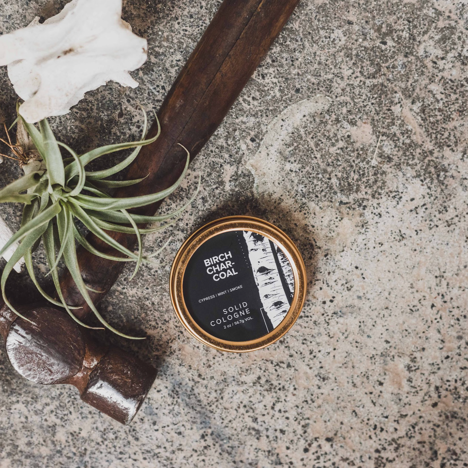 Birch Charcoal solid cologne on concrete next to hammer and green floral