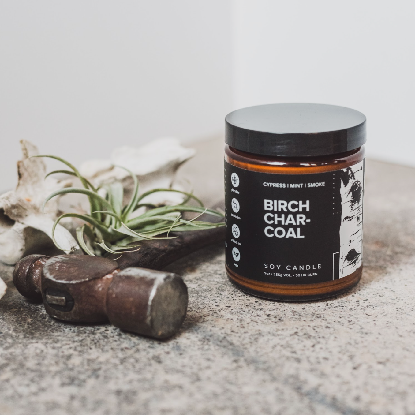 9oz Birch Charcoal candle on cement next to a hammer and green foliage