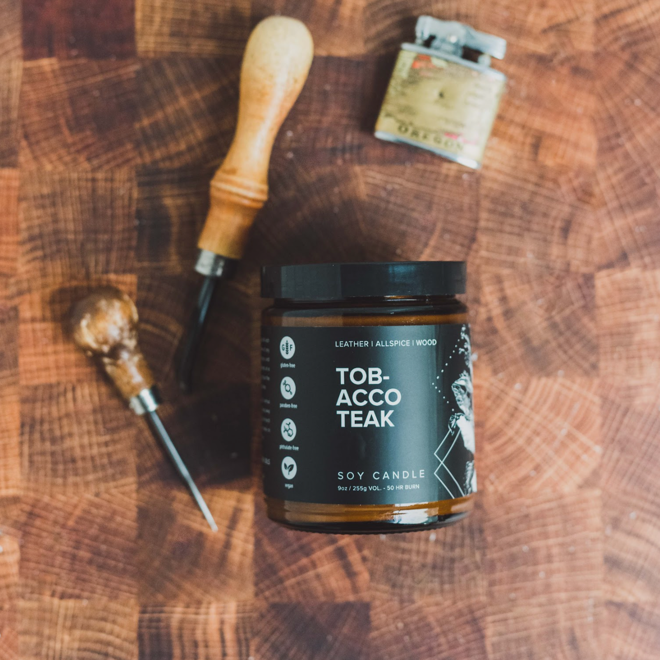 9oz Tobacco Teak candle on wood table next to tools and lighter