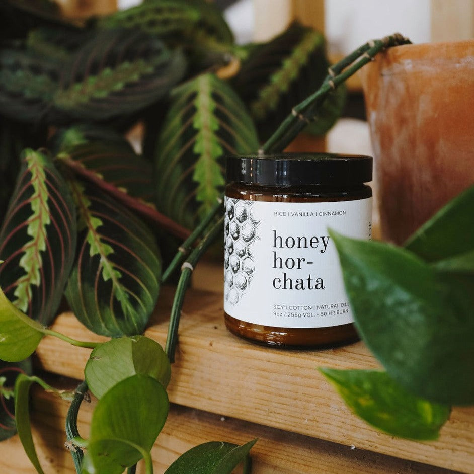 9oz Honey Horchata candle on wooden shelf and green foliage