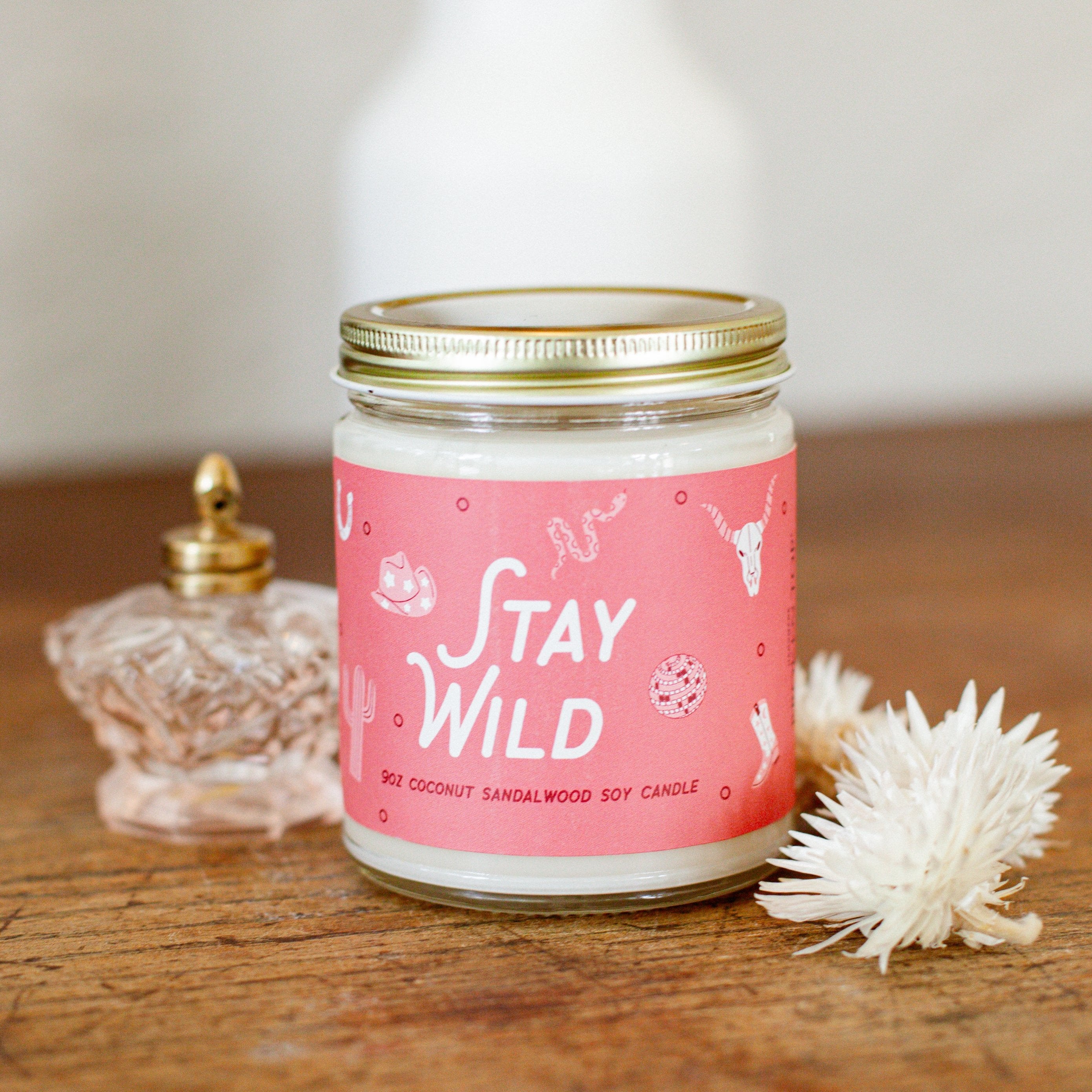 9oz 'Stay Wild' Soy Candle on a wooden table next to decorative glass jar and flowers. 