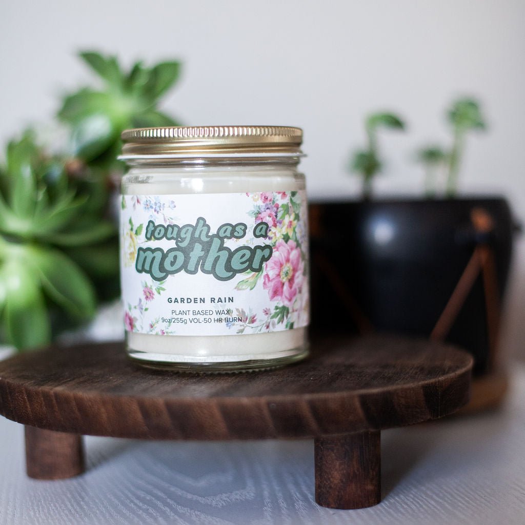 9oz 'Tough As A Mother' Soy Candle on wooden stand on white table with potted plants in background