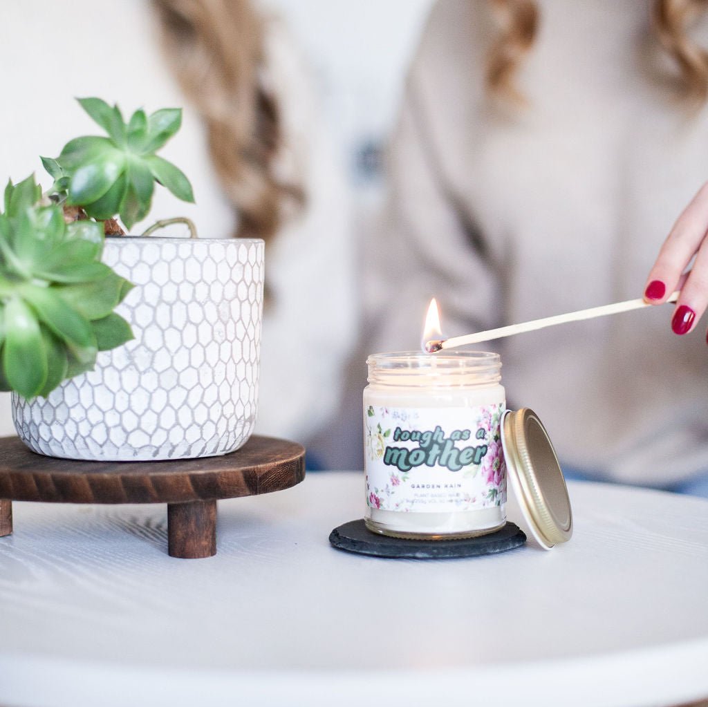 Two women lighting 9oz 'Tough As A Mother' Soy Candle on white table next to potted plant