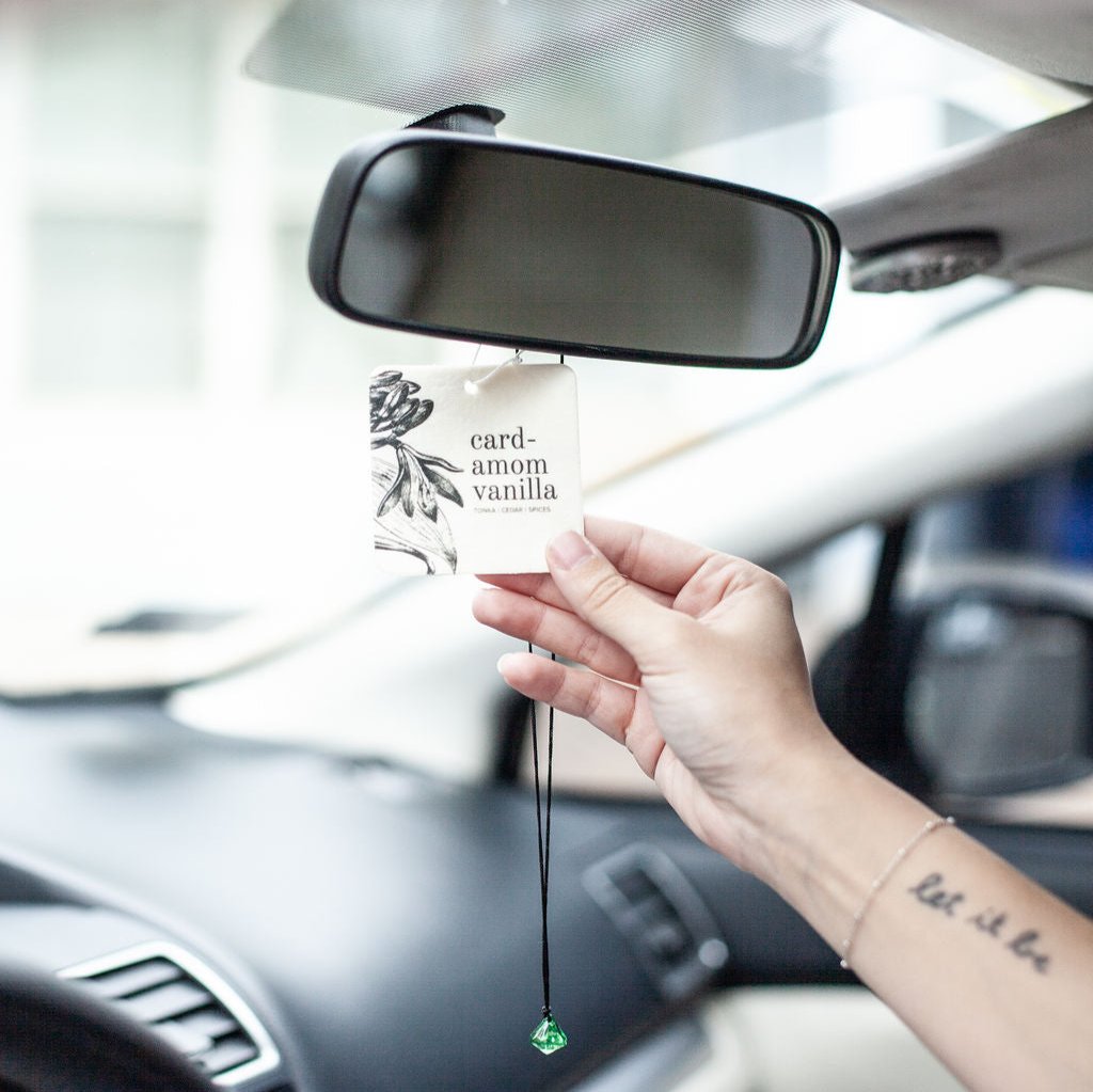 Cardamom Vanilla Car Freshener hanging from rear view mirror with hand grasping it. 