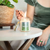 Young woman holding lit match about to light 9oz 'Daydreamer' Soy Candle on white table