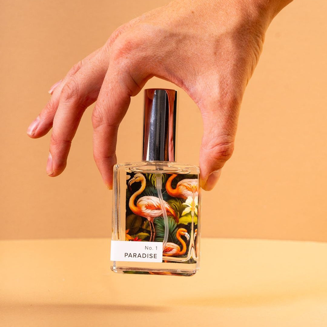 Hand holding a perfume bottle labeled 'Paradise' with a colorful design on a beige background