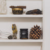 9oz Santal Noir candle on white shelf next to pine cone and other home decor