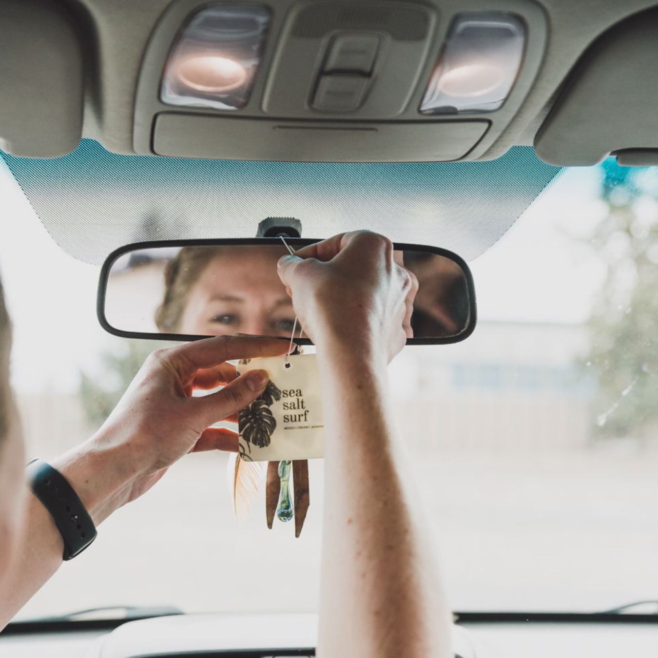 Sea Salt Surf Car Freshener being hung on a rearview mirror by two hands. 