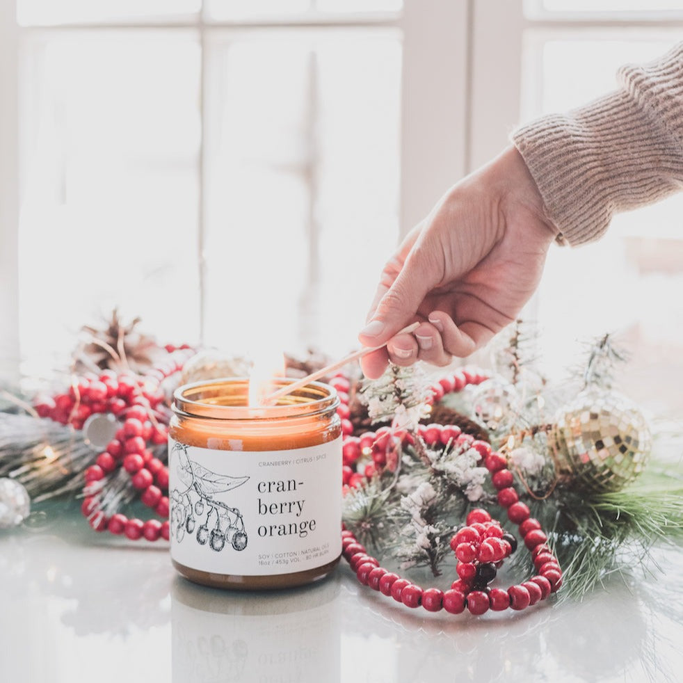 16oz Cranberry Orange candle on glass table being lit by women's hand and holiday decor